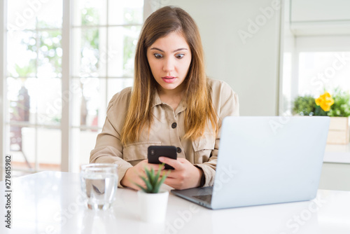 Beautiful young woman using smartphone and computer scared in shock with a surprise face  afraid and excited with fear expression