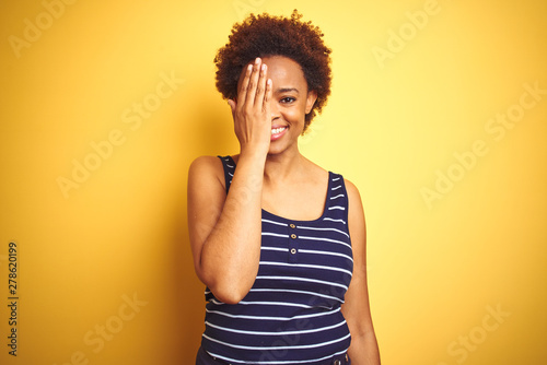 Beauitul african american woman wearing summer t-shirt over isolated yellow background covering one eye with hand, confident smile on face and surprise emotion.