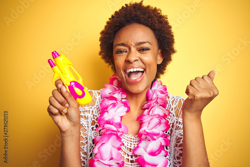 Young african american woman with afro hair wearing flower hawaiian lei and water gun screaming proud and celebrating victory and success very excited, cheering emotion