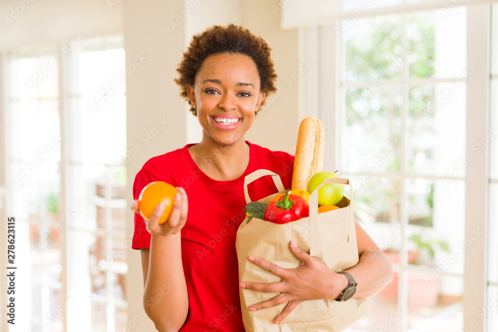 Young beautiful african american woman holding paper bag full of fresh healthy groceries and picking vegetables Young beautiful african american woman holding paper bag full of fresh healthy groceries and picking vegetables