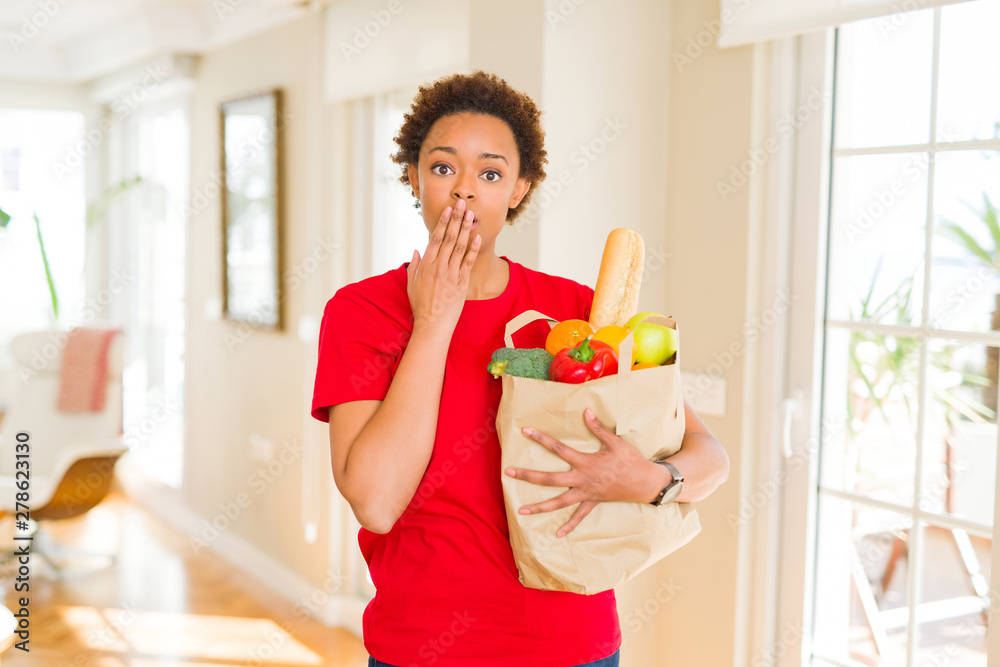 Young african american woman holding paper bag full of fresh groceries cover mouth with hand shocked with shame for mistake, expression of fear, scared in silence, secret concept Young african american woman holding paper bag full of fresh groceries cover mouth with hand shocked with shame for mistake, expression of fear, scared in silence, secret concept