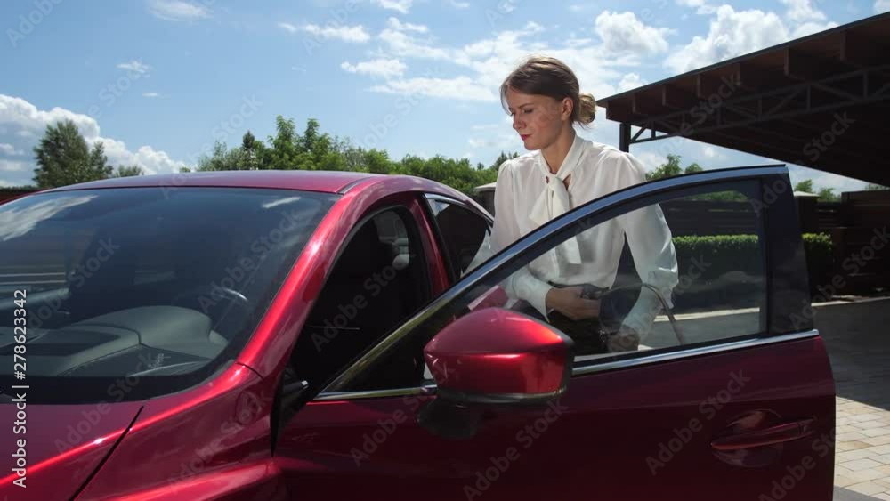 Close-up of adult female in formal clothes getting into car going on business, bright sun reflected in windshield. Confident woman driving to office by own auto standing on courtyard path near house