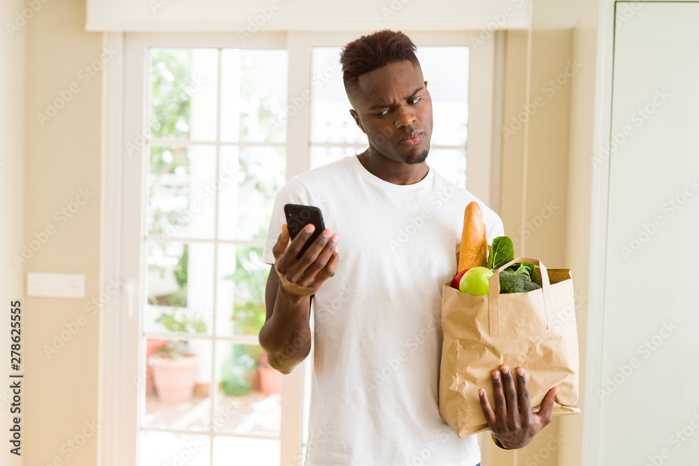 African man holding a paper bag full of groceries and using smarpthone buying online using app African man holding a paper bag full of groceries and using smarpthone buying online using app