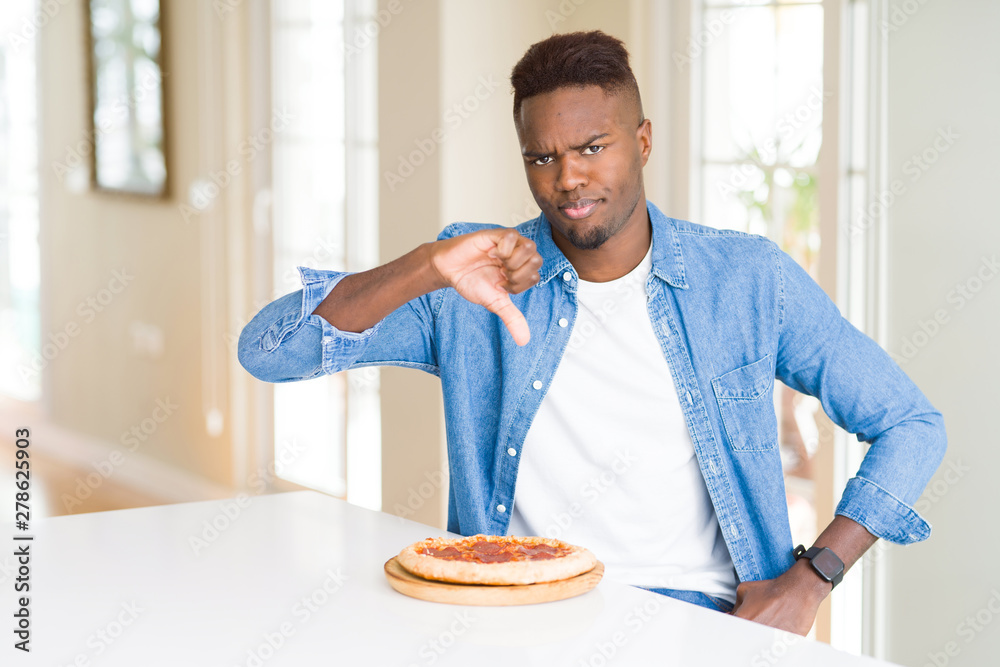 African american man eating pepperoni pizza at home with angry face, negative sign showing dislike with thumbs down, rejection concept African american man eating pepperoni pizza at home with angry face, negative sign showing dislike with thumbs down, rejection concept