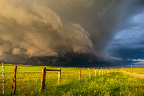 A dark shelf cloud and storm approach as the sun shines brightly looking down a fence in the rural countryside.