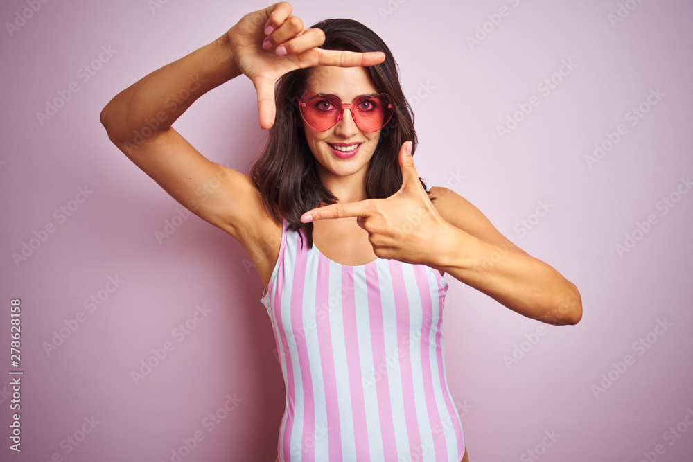 Beautiful woman wearing striped pink swimsuit and heart shaped sunglasses over pink background smiling making frame with hands and fingers with happy face. Creativity and photography concept. Beautiful woman wearing striped pink swimsuit and heart shaped sunglasses over pink background smiling making frame with hands and fingers with happy face. Creativity and photography concept.