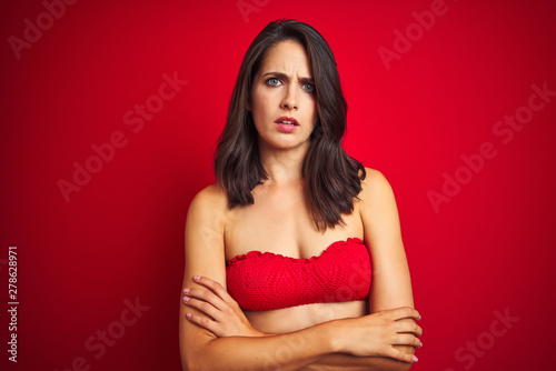 Beautiful woman wearing bikini swimwear over red isolated background skeptic and nervous, disapproving expression on face with crossed arms. Negative person.