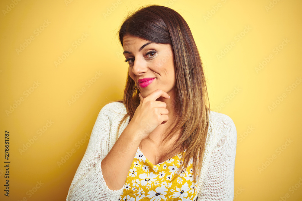 Young beautiful woman wearing jacket standing over yellow isolated background looking confident at the camera smiling with crossed arms and hand raised on chin. Thinking positive.