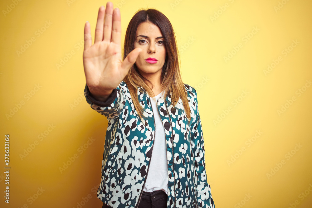 Young beautiful woman wearing casual jacket over yellow isolated background doing stop sing with palm of the hand. Warning expression with negative and serious gesture on the face. Young beautiful woman wearing casual jacket over yellow isolated background doing stop sing with palm of the hand. Warning expression with negative and serious gesture on the face.