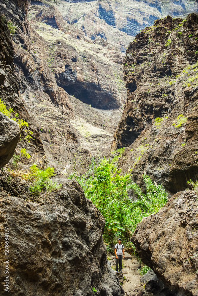 Young traveler stays on the top of huge boulder in the Masca gorge, Tenerife, showing solidified volcanic lava flow layers and arch formation. Ravine leads down to the ocean from a 900m altitude