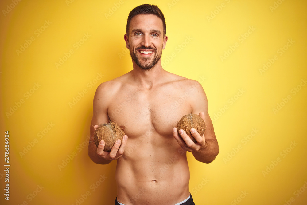 Young handsome shirtless man holding exotic tropical coconut over isolated yellow background with a happy face standing and smiling with a confident smile showing teeth