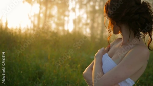 A beautiful girl in a white dress is alone in the summer forest, looking toward the dawn of the sun, hugging her arms. The average plan.