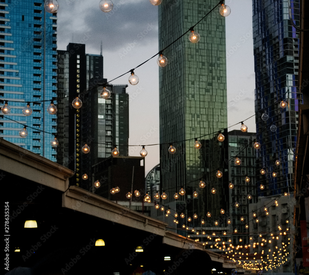 Fototapeta premium A hanging string of lights at Queen Victoria Markets with the skyline of Melbourne city in the background