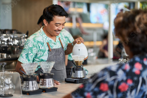 Candid lifestyle shot of smiling ethnic indonesian barista and small business owner preparing organic fair-trade coffee in bright trendy coffee shop wearing apron photo
