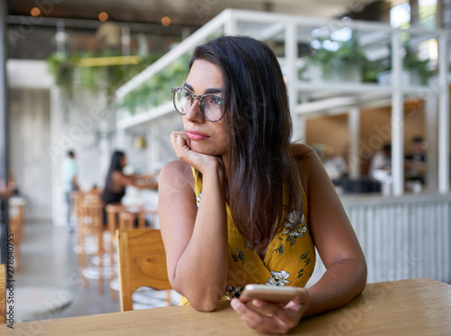 Candid lifestyle portrait of beautiful hispanic millennial woman looking out window holding cellphone in modern trendy and bright coffee shop with plants photo