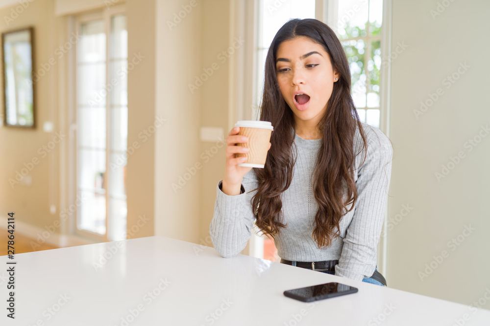 Young woman drinking a cup of coffee at home scared in shock with a surprise face, afraid and excited with fear expression