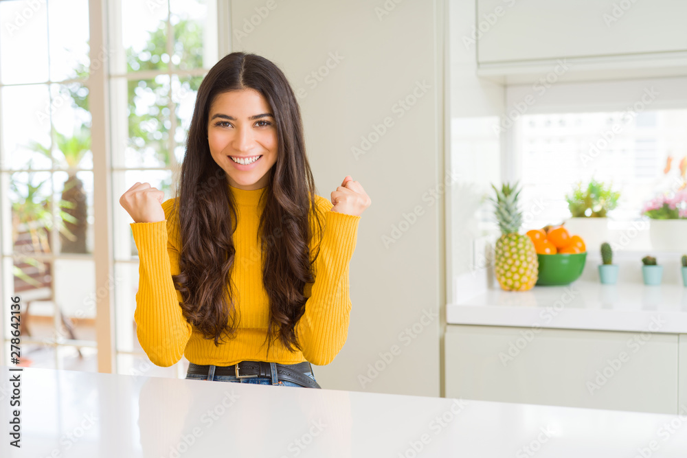 Young beautiful woman at home on white table celebrating surprised and amazed for success with arms raised and open eyes. Winner concept. Young beautiful woman at home on white table celebrating surprised and amazed for success with arms raised and open eyes. Winner concept.