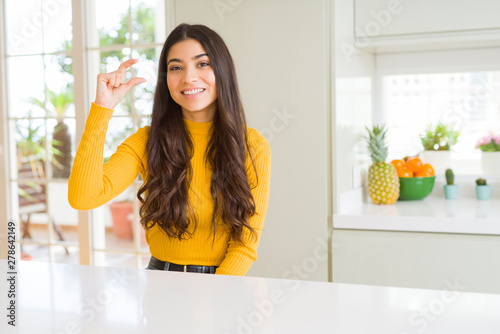 Young beautiful woman at home on white table smiling and confident gesturing with hand doing size sign with fingers while looking and the camera. Measure concept.