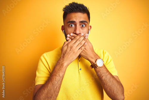 Young indian man listening to music using earphones standing over isolated yellow background shocked covering mouth with hands for mistake. Secret concept.