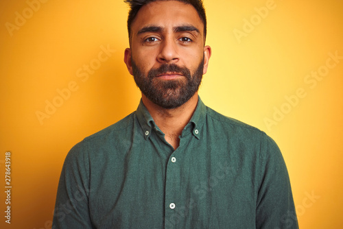 Young indian man wearing green shirt standing over isolated yellow background with serious expression on face. Simple and natural looking at the camera. © Krakenimages.com
