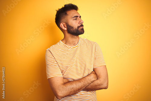 Young indian man wearing t-shirt standing over isolated yellow background looking to the side with arms crossed convinced and confident