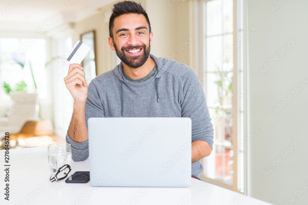 Handsome man smiling using credit card as payment metod when shopping online using laptop
