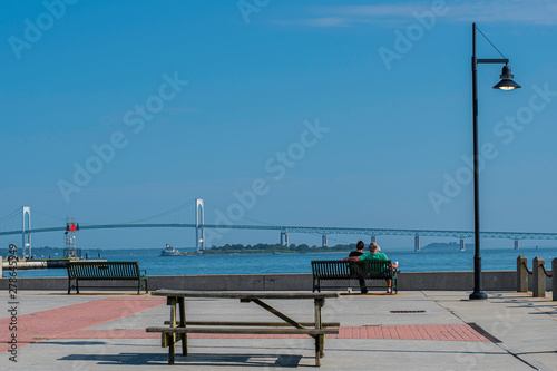 Man in green shirt and woman in black sitting on green bench near water with coffee cup watching boats and looking at the bridge.