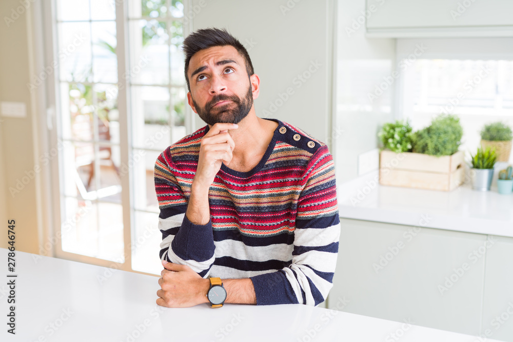 Handsome hispanic man wearing stripes sweater at home with hand on chin thinking about question, pensive expression. Smiling with thoughtful face. Doubt concept. Handsome hispanic man wearing stripes sweater at home with hand on chin thinking about question, pensive expression. Smiling with thoughtful face. Doubt concept.