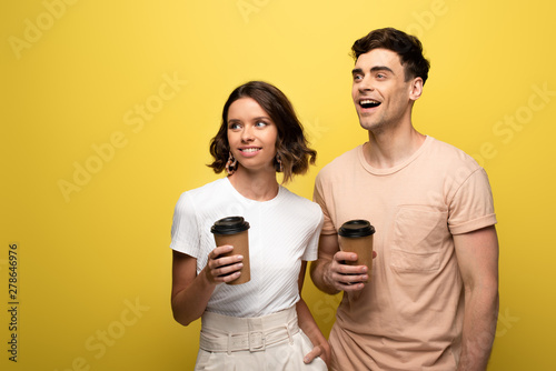 cheerful man and woman holding disposable cups while looking away on yellow background