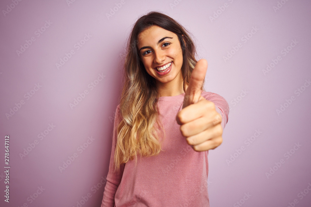 Young beautiful woman wearing a sweater over pink isolated background doing happy thumbs up gesture with hand. Approving expression looking at the camera showing success. Young beautiful woman wearing a sweater over pink isolated background doing happy thumbs up gesture with hand. Approving expression looking at the camera showing success.