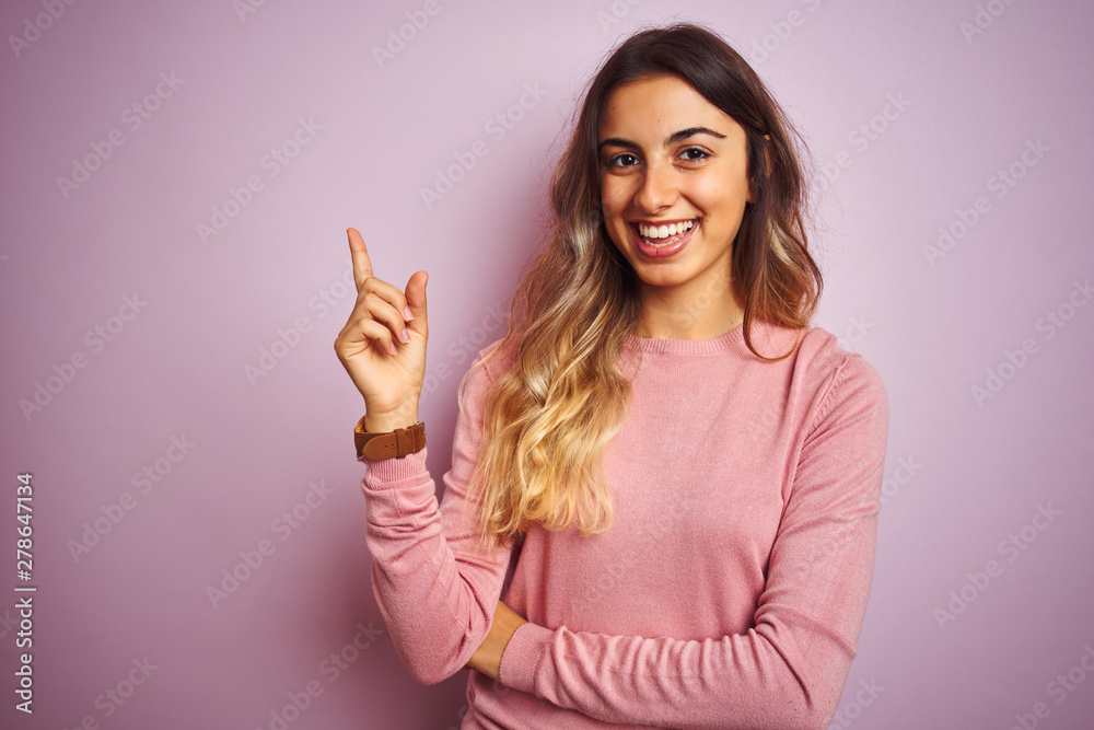 Young beautiful woman wearing a sweater over pink isolated background with a big smile on face, pointing with hand and finger to the side looking at the camera. Young beautiful woman wearing a sweater over pink isolated background with a big smile on face, pointing with hand and finger to the side looking at the camera.