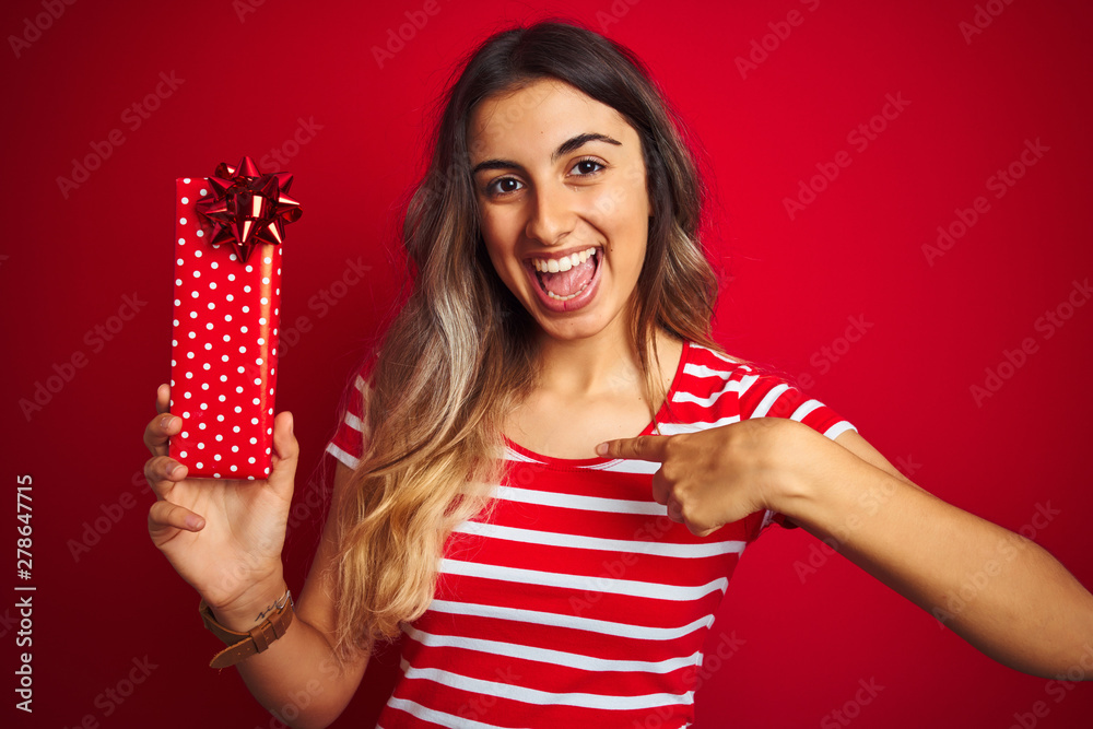 Young beautiful woman holding birthday present over red isolated background with surprise face pointing finger to himself Young beautiful woman holding birthday present over red isolated background with surprise face pointing finger to himself