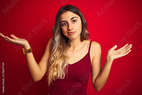 Young beautiful woman wearing a t-shirt over red isolated background clueless and confused expression with arms and hands raised. Doubt concept. © Krakenimages.com