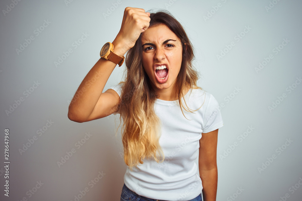 Young beautiful woman wearing casual white t-shirt over isolated background angry and mad raising fist frustrated and furious while shouting with anger. Rage and aggressive concept. Young beautiful woman wearing casual white t-shirt over isolated background angry and mad raising fist frustrated and furious while shouting with anger. Rage and aggressive concept.