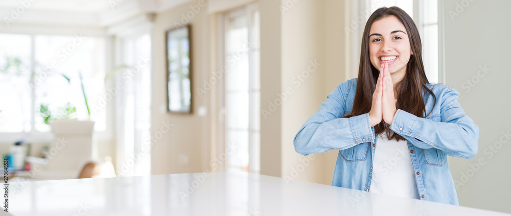 Wide angle picture of beautiful young woman sitting on white table at home praying with hands together asking for forgiveness smiling confident. Wide angle picture of beautiful young woman sitting on white table at home praying with hands together asking for forgiveness smiling confident.