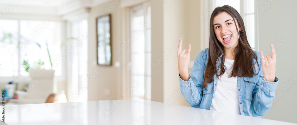Wide angle picture of beautiful young woman sitting on white table at home shouting with crazy expression doing rock symbol with hands up. Music star. Heavy concept.
