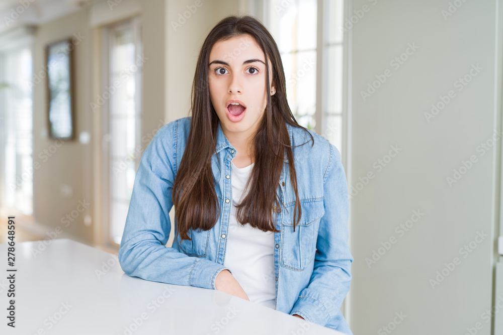 Beautiful young woman sitting on white table at home afraid and shocked with surprise expression, fear and excited face. Beautiful young woman sitting on white table at home afraid and shocked with surprise expression, fear and excited face.