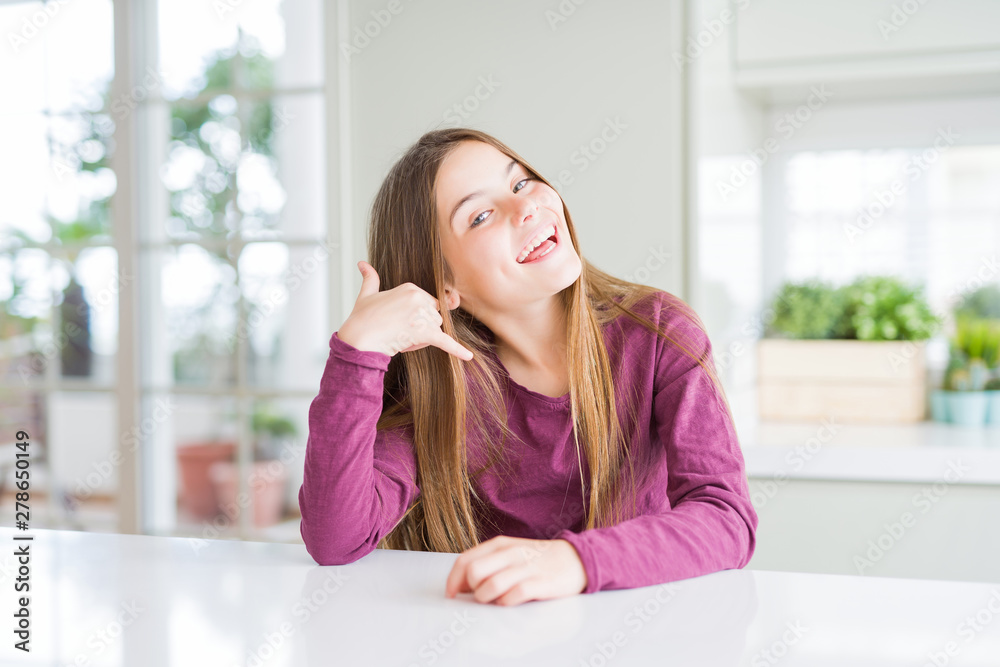 Beautiful young girl kid on white table smiling doing phone gesture with hand and fingers like talking on the telephone. Communicating concepts.