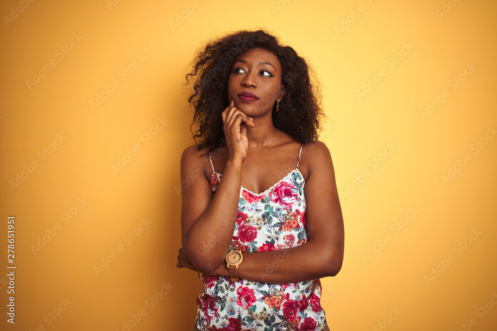 African american woman wearing floral summer t-shirt over isolated yellow background with hand on chin thinking about question, pensive expression. Smiling with thoughtful face. Doubt concept. African american woman wearing floral summer t-shirt over isolated yellow background with hand on chin thinking about question, pensive expression. Smiling with thoughtful face. Doubt concept.