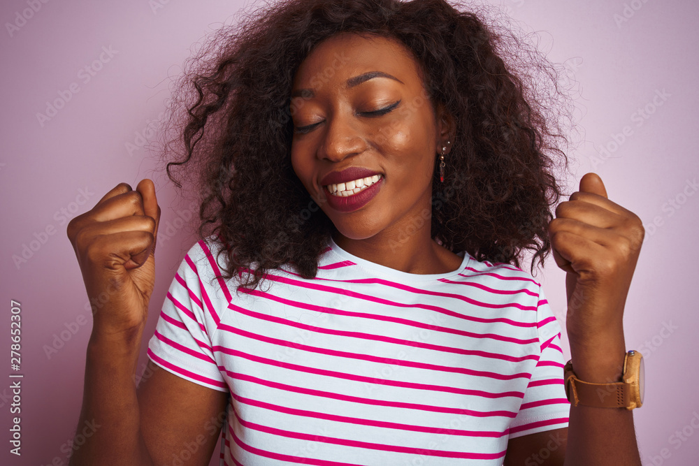 Young african american woman wearing striped t-shirt standing over isolated pink background very happy and excited doing winner gesture with arms raised, smiling and screaming for success. 