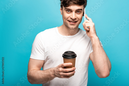 positive young man looking at camera while talking on smartphone and holding paper cup on blue background