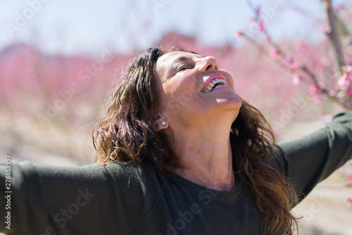 Beautiful middle age woman in the middle of pink peach flowers and trees smiling cheerful with open arms enjoying sunbathe on sunny day