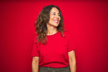 Middle age senior woman with curly hair over red isolated background looking away to side with smile on face, natural expression. Laughing confident.