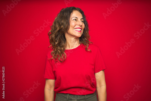 Middle age senior woman with curly hair over red isolated background looking away to side with smile on face, natural expression. Laughing confident.