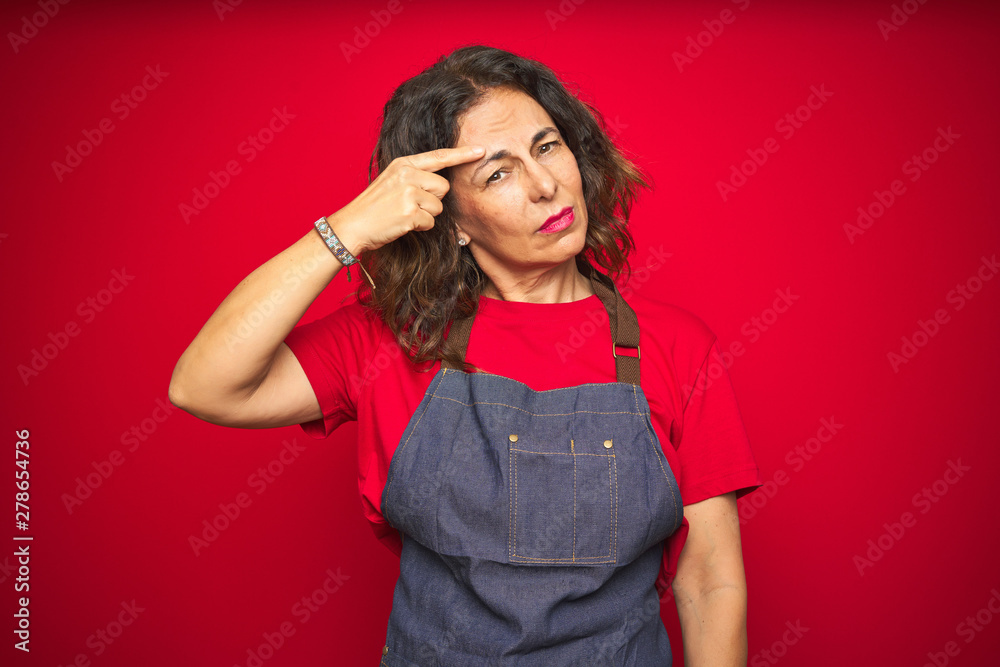Middle age senior woman wearing apron uniform over red isolated background pointing unhappy to pimple on forehead, ugly infection of blackhead. Acne and skin problem Middle age senior woman wearing apron uniform over red isolated background pointing unhappy to pimple on forehead, ugly infection of blackhead. Acne and skin problem