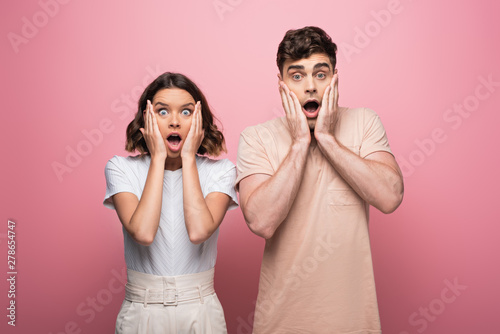 shocked man and woman holding hands near face while looking at camera on pink background