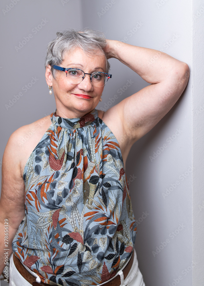 Elderly Woman with short gray hair and glasses posing in front of light gray  wall wearing