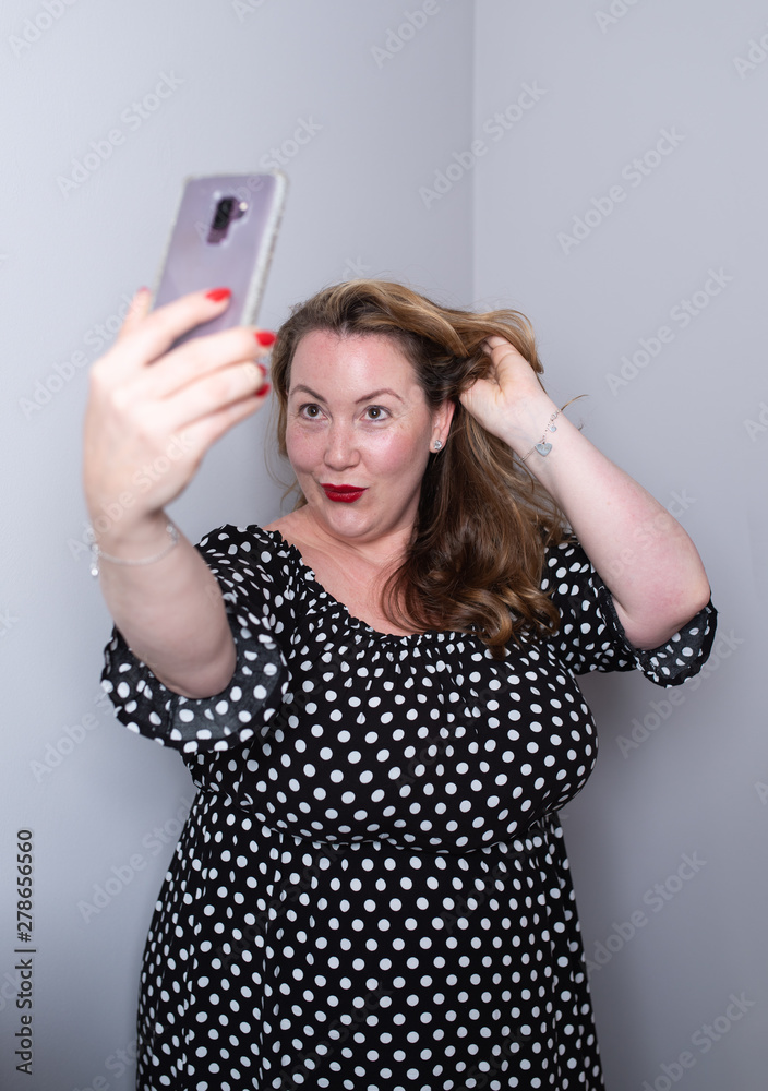 Beautiful overweight plus size model taking selfies for social media with her smart phone. the woman is touching her hair and making a duckface wearing a black dress with white dots Beautiful overweight plus size model taking selfies for social media with her smart phone. the woman is touching her hair and making a duckface wearing a black dress with white dots