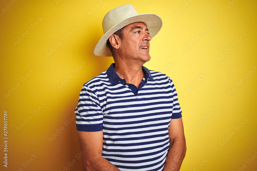Handsome middle age man wearing striped polo and hat over isolated yellow background looking away to side with smile on face, natural expression. Laughing confident.