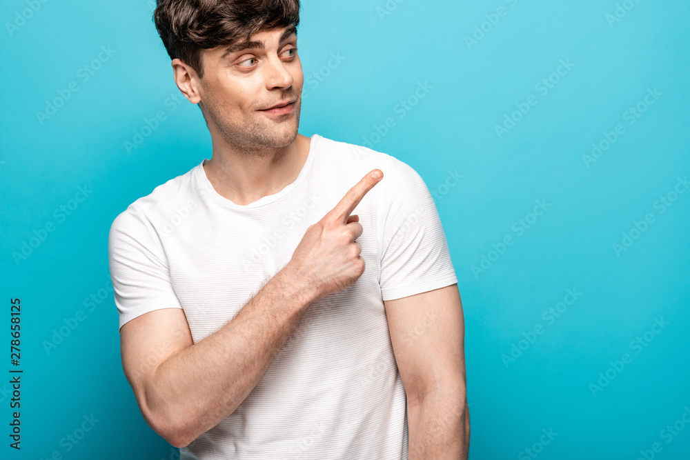 handsome young man smiling while looking away and pointing with finger on blue background handsome young man smiling while looking away and pointing with finger on blue background